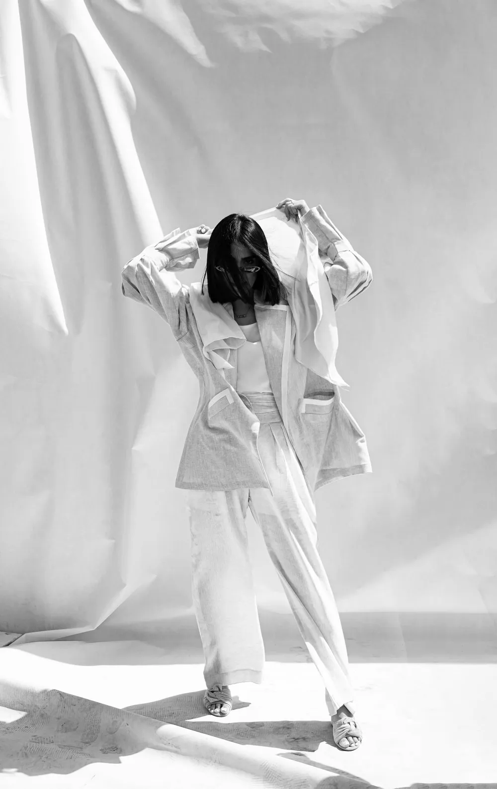 Mujer con conjunto blanco de saco y pantalón, los brazos sobre la cabeza, entre telas claras