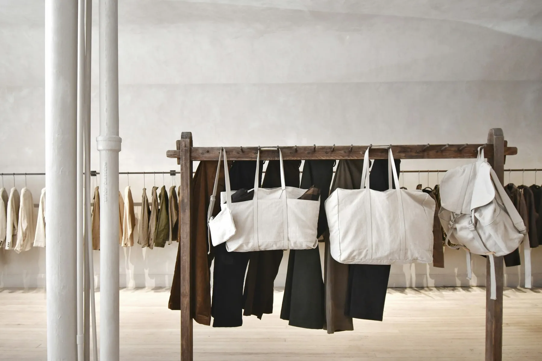 Perchero de madera con prendas y bolsos de lona colgados en un salón amplio de paredes claras y piso de madera